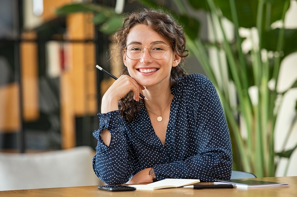 Young Woman Wearing Glasses and Smiling at Desk – Glasses Exams A cheerful woman wearing stylish eyeglasses sits at her workspace, enjoying clear and comfortable vision. Regular glasses exams help maintain sharp eyesight and keep prescriptions up to date – Glasses Exams.