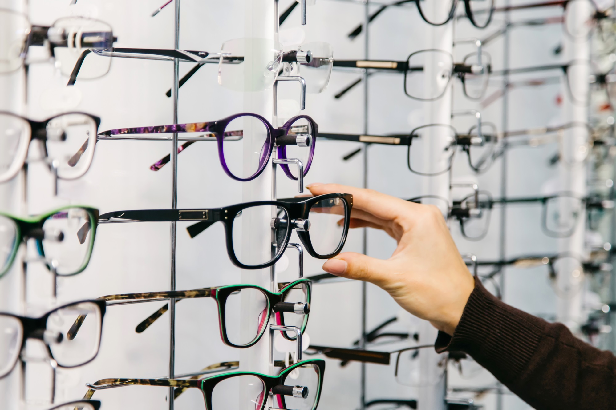 A hand reaches toward a stylish black frame on a display wall filled with various eyeglass styles. The image highlights the wide selection of fashion-forward frames available for every preference – Eyeglasses.