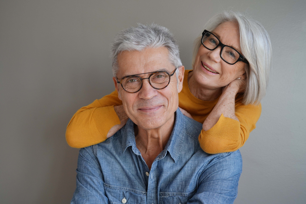A cheerful senior couple stands closely together, both wearing stylish glasses that help them maintain clear vision. Their warm expressions show how proper eye care and corrective lenses can significantly improve quality of life for those dealing with blurry vision.