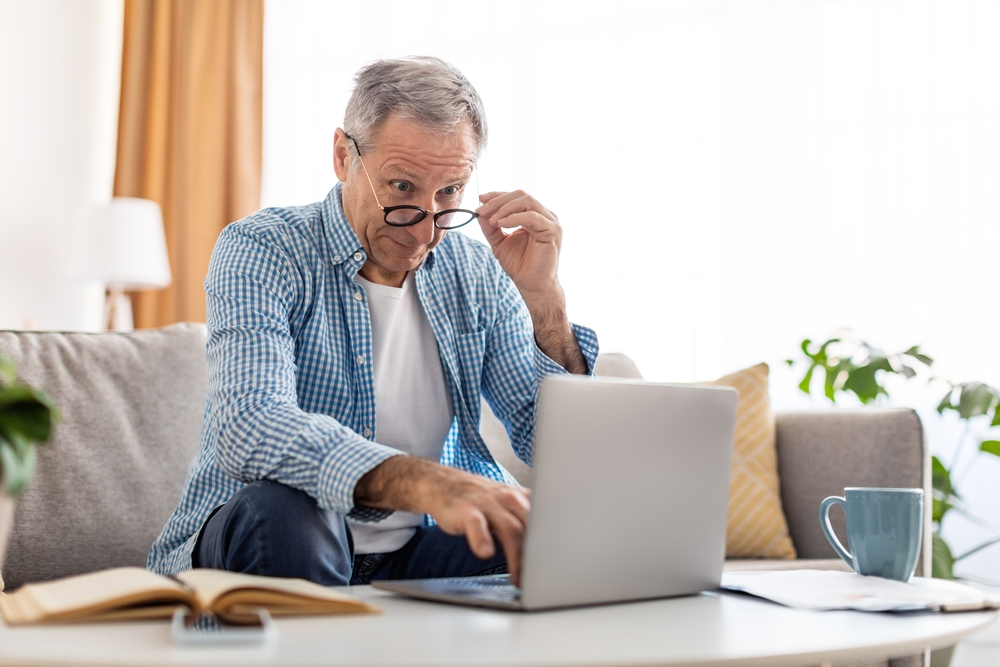 An older man sits on his couch, leaning in toward his laptop as he adjusts his glasses to see more clearly. His posture and expression reflect the common struggle of blurry vision that often disrupts daily activities like reading or working on a computer.